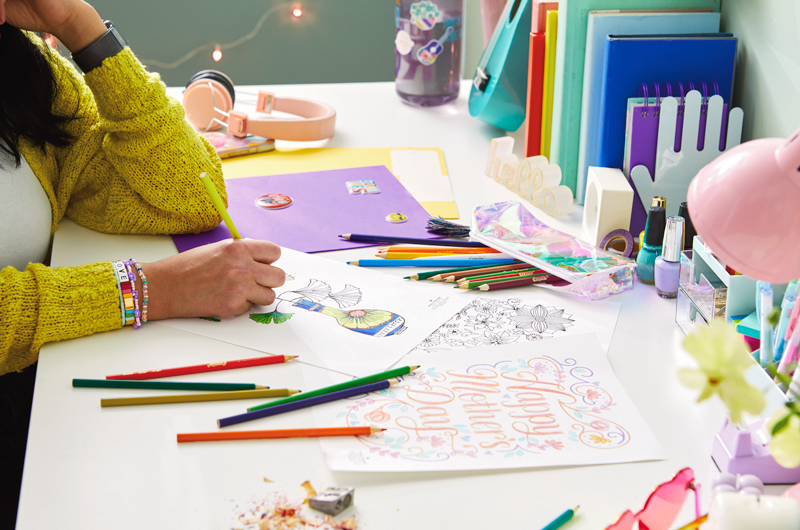 A woman in a bright yellow sweater sitting at a desk and coloring an intricately designed free printable Mother's Day coloring page.