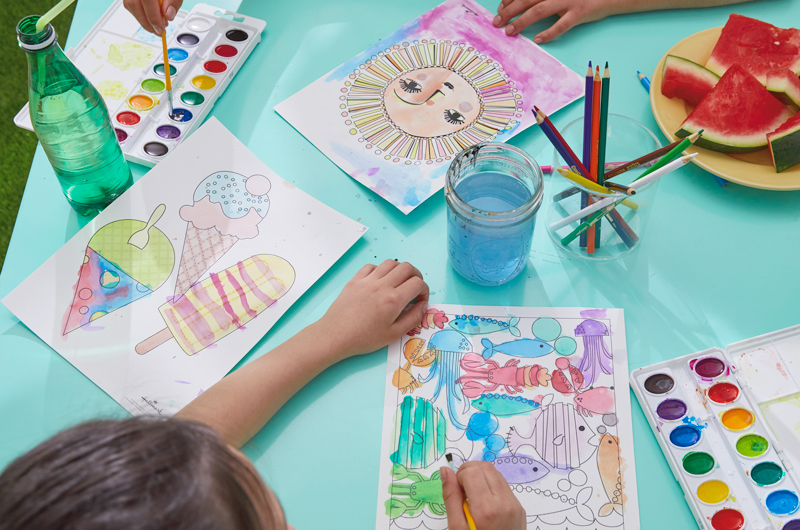 Two children sit at an aqua colored table with free printable summer coloring pages, slices of watermelon, watercolor paints and colored pencils.