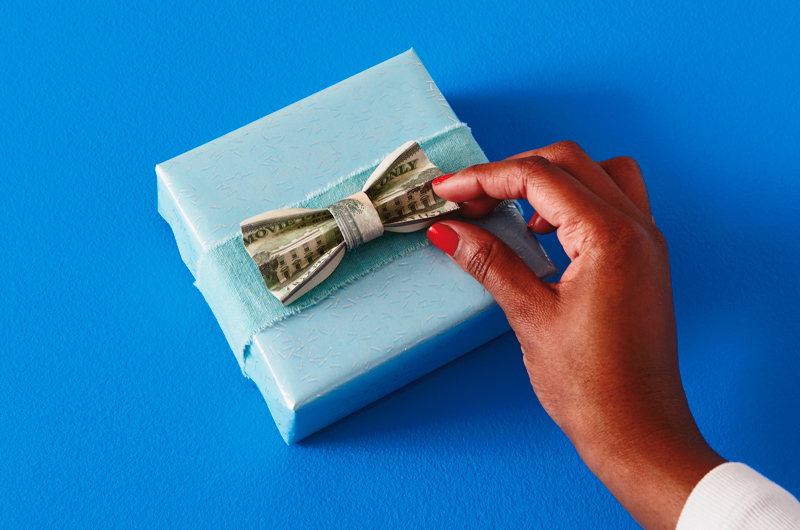 A woman's hand reaches into frame to adjust a dollar bill that's been folded accordian style and wrapped with another dollar bill to look like a bow tie. The cash bow tie sits atop a gift wrapped in light blue wrapping paper.