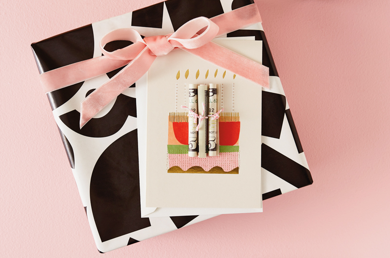Five dollar bills are tightly rolled up individually and tied with small pieces of string, then attached to a card depicting a cake and candles so that the rolled up bills look like candles. The card sits on top of a gift wrapped with black and white graphic wrapping paper and tied with a pink felt ribbon.