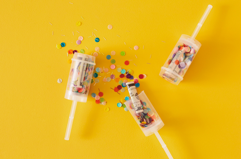 Clear DIY confetti party poppers filled with confetti and rolled up dollar bills are spilling out onto a bright yellow surface.