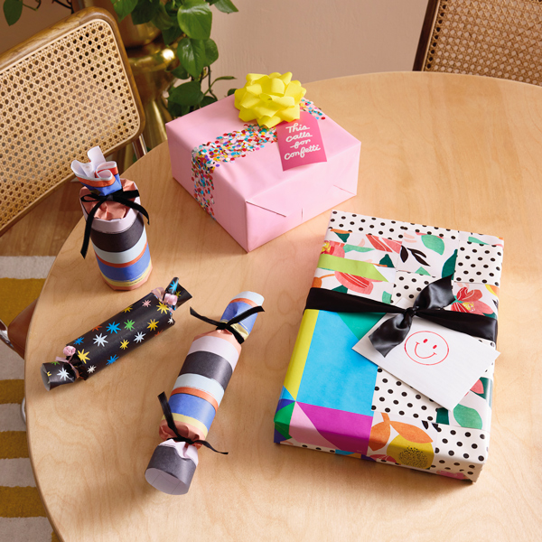 Gifts sitting on a table after being wrapped using creative ways to use gift wrap scraps—one making a quilt pattern out of leftover pieces, another with a wrapping paper confetti accent band, and the final one made from the cardboard tube that goes inside a roll of gift wrap, wrapped and tied at the ends like a piece of candy.