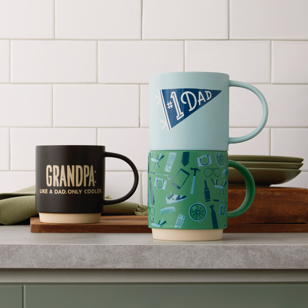 A selection of dad mugs on a granite countertop with a white subway tile backsplash. Two of the mugs read, 