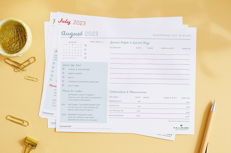 A free printable August celebration planner page sits on a light yellow surface with a scattering of gold paper clips and a pen nearby.