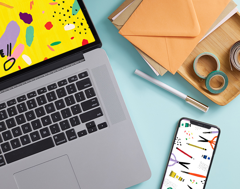 A laptop is open and shows a digital wallpaper with a bright yellow background and various bright colored shapes as its digital background; lying right next to it is a mobile phone with a digital background that features illustrations of art supplies like paint tubes, scissors, pencils, paint brushes, and paper scraps.