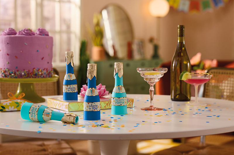 A table covered in birthday party trimmings, including confetti poppers, a cake with purple icing and sprinkles on a cake stand, wrapped gifts with bows on top, cocktail glasses and a wine bottle.