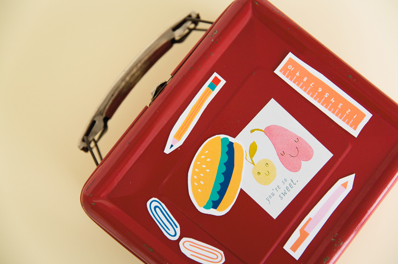 Our free, DIY back-to-school magnets clinging to the side of a red metal lunchbox, holding a note that features an illustration of two pieces of fruit and the message, 