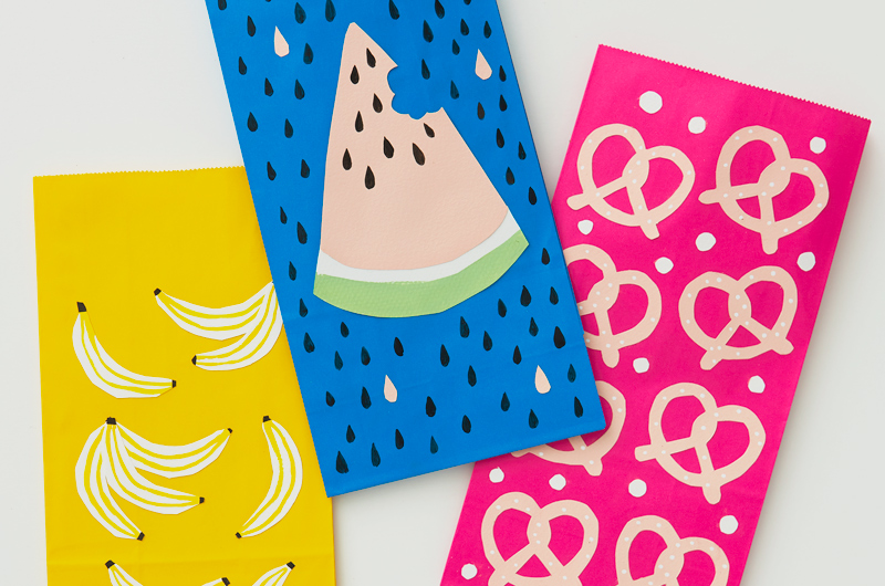 Three brightly colored paper lunch sacks are covered in cut-paper in the shapes of different snack foods, including bananas, watermelon and pretzels.