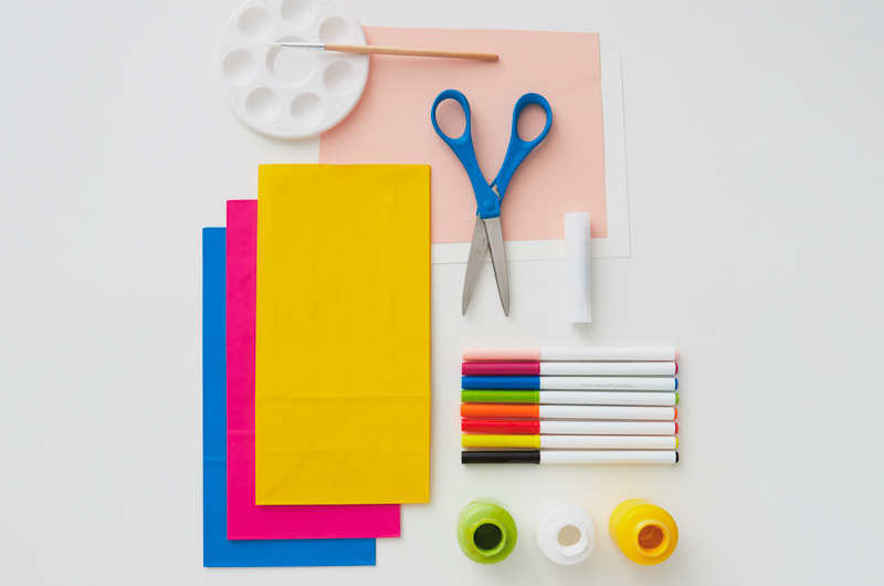 Supplies needed for this DIY are paper lunch sacks, construction paper, scissors, a glue stick, a paint palette and brush, and paints.
