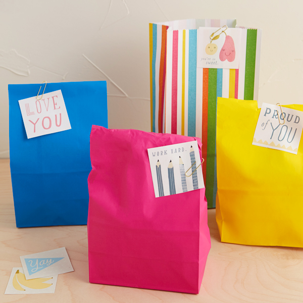 Four brightly colored paper lunch sacks sit on a white table top, each with one of our free, printable lunch box notes taped to it.
