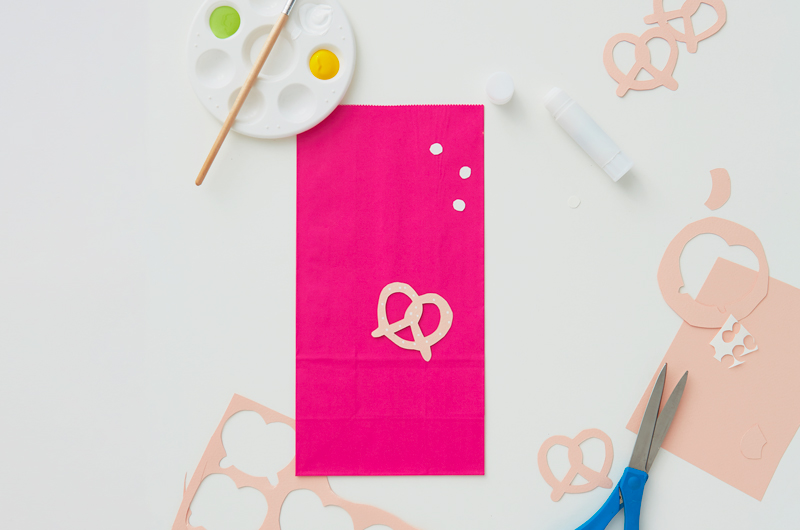 A bright pink paper lunch sack lays on a white surface with light pink paper cut into the shape of pretzels strewn around; a pair of scissors, a glue stick, and a paint palette and brush lay nearby.