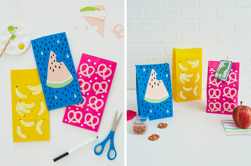 Three paper lunch sacks—a blue one, a yellow one, and a bright pink one—are decorated with cut paper shapes of different snacks, including watermelon, bananas and pretzels; they're sitting on a white countertop in front of a white brick background, with a container of mini pretzels, an apple, and a Just Because mini card sitting nearby.