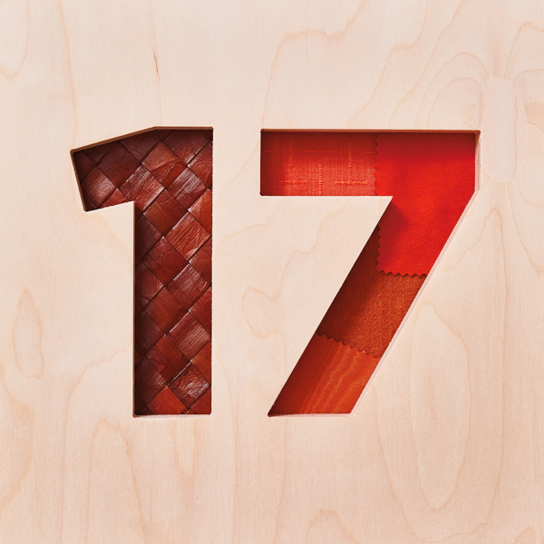 The number 17 has been cut out of a piece of plywood, with woven caning and fabric patches showing through underneath the cutout numbers, symbolizing the traditional 17th wedding anniversary gift of furniture.