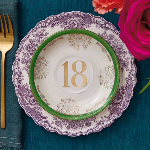 A china bowl with a green and gold rim since on top of a china plate with a scalloped rim and a dark purple rose pattern; at the bottom of the bowl is the number 18 in gold metallic lettering, symbolizing the traditional 18th wedding anniversary gift of china.