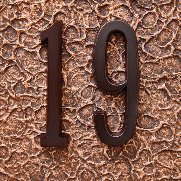 The number 19 is wrought in bronze-colored metal and sits atop a bronze-colored, metallic, wavy surface, symbolizing the traditional 19th wedding anniversary gift, bronze.