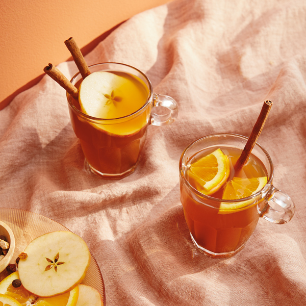 Two clear glass mugs of warm mulled cider, garnished with apple slices, orange slices and cinnamon sticks.