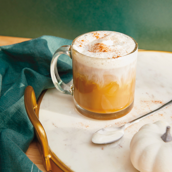 A pumpkin spice latte in a clear glass mug sits on a white marble serving tray with gold handles.