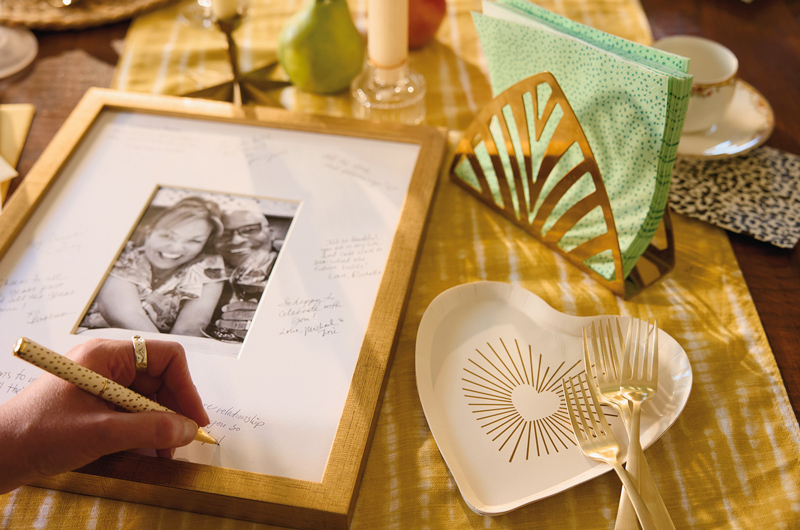 A woman's hand signs the mat of a framed photo of the anniversary couple with a gold polka-dotted pen; next to the gold framed photo is a white and gold, heart-shaped Celebrate! dessert plate, as well as a vintage napkin holder filled with aqua and teal polka dotted Celebrate! napkins.