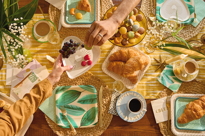 A downshot of a party table set up, laid with varying patterns of Celebrate! partyware topped with breakfast foods like croissants, berries and macarons, as well as cups of coffee in vintage cups and saucers.