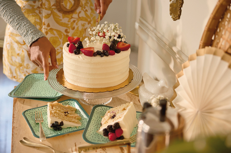A cake with white icing and topped with berries and baby's breath sits on a clear glass cake stand, surrounded by teal polka dotted Celebrate! dessert plates; a woman stands behind the cake, preparing to cut it.