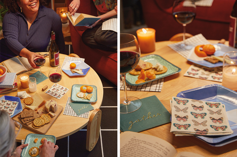 A book club party is set up on a low table, with Celebrate! partyware in colors of blue and green, cheese and crackers, fruit and glasses of wine; a woman sitting on the floor next to the table smiles up at her friend who is holding an open book, curled up on the couch nearby.