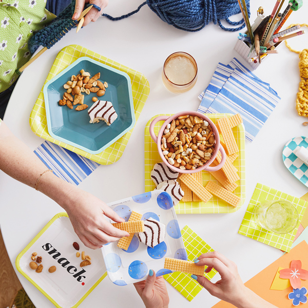 A downshot of a bright, colorful craft party table setup; a woman reaches across the table to offer a plate of snacks to another woman who is knitting on the other side of the table.