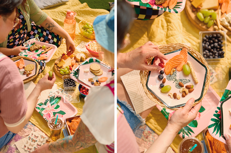 A thrift store picnic party is set up on a quilted yellow blanket, which is laden with Celebrate! partyware in floral and butterlfy patterns and finger foods like fruit, crackers and cheese.
