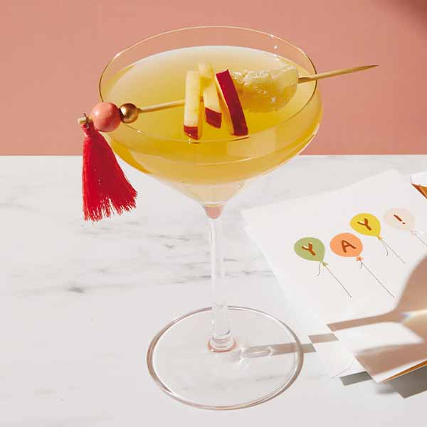 A spiced appletini in a couple glass is garnished with apple slices and crystalized ginger that has been speared onto a cocktail stick with a red tassel on one end; lying at the foot of the glass is a greeting card with lettered balloons that spell out 