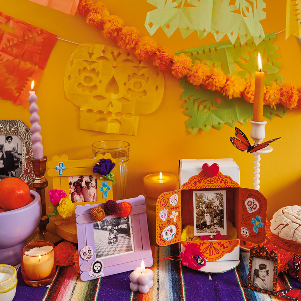 A Dia de Muertos ofrenda is decorated with DIY papel picado and DIY Day of the Dead crafts for kids, including a DIY shadowbox and a DIY ofrenda photo frame made of painted popsicle sticks.