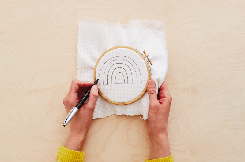 A woman's hands sketch a rainbow pattern onto a piece of evenweave fabric that's been fused with interfacing and inserted into an embroidery hoop.