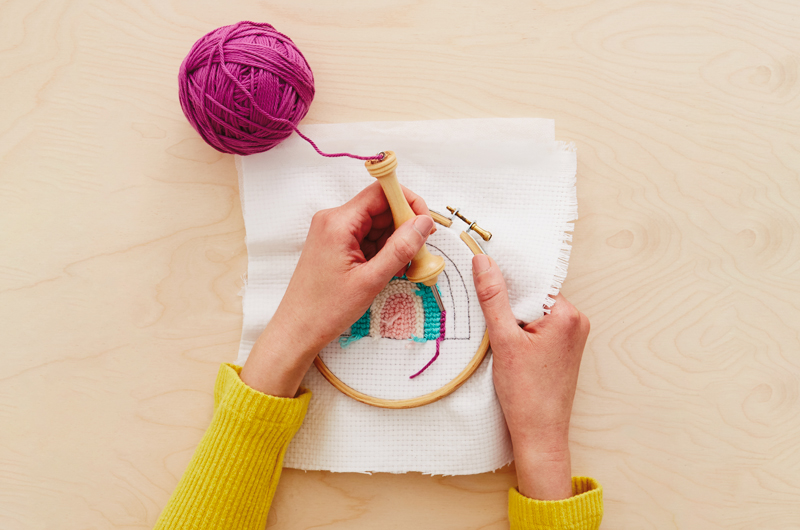 A woman uses a punch needle to create a rainbow pattern in a piece of evenweave fabric that's been inserted into an embroidery hoop; the first three inner arches of the rainbow have been embroidered with light pink, white and aqua yarn, and she is starting a fourth arch with magenta colored yarn.