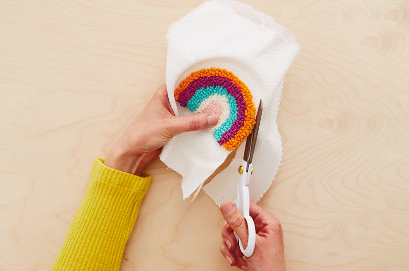 A woman uses a pair of scissors to trim excess fabric from her completed needle punch patch in the shape of a rainbow.