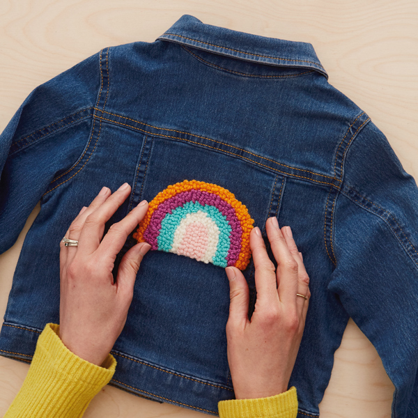 A woman's hands center a needle punch patch in the shape of a brightly colored rainbow onto the back of a dark blue denim jacket.
