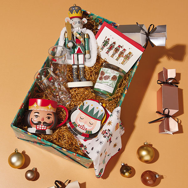 A Christmas care package filled with nutcracker-themed gifts, including nesting snack bowls, juice glasses, a mug, a mouse king nutcracker, a tea towel with a nutcracker pattern printed on it, and a nutcracker holiday greeting card.