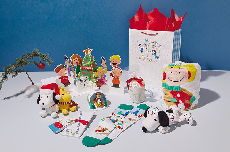 Gifts for a Peanuts-themed Christmas care package include a Snoopy and Woodstock Better Together magnetic plush set, a floppy Snoopy covered in a Christmas light pattern, cozy socks featuring a winter sledding scene with all the Peanuts Gang members, a cozy fleece blanket featuring the Peanuts gang, a ceramic mug featuring a line illustration of Charlie Brown and Snoopy ice skating together, a pop-up Peanuts card, a dimensional desk figurine featuring Charlie Brown and Snoopy sledding down a wooded, snow-covered hill, and a large gift bag with an illustration of Charlie Brown, Lucy, Snoopy and Woodstock building snowmen.