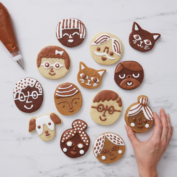 A variety of cookies decorated to look like the faces of different people and pets and a piping bag full of chocolate icing lay on a white marble countertop; a woman's hand is reaching into frame to take one.