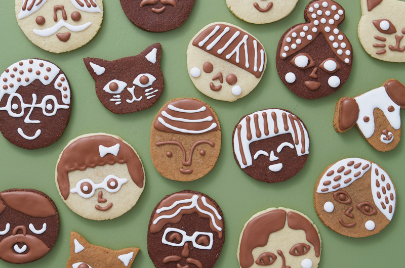 A variety of cookies decorated to look like different people's and pet's faces sit on a light green surface; the cookies appear in three different flavors—chocolate, sugar cookie and gingerbread—and are decorated with white and chocolate royal icing.