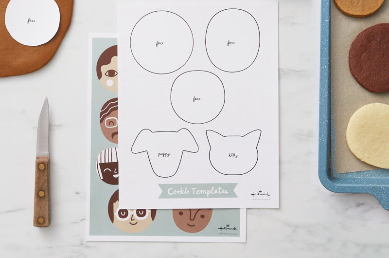 A free cookie face template printable sits on a white marble countertop with an example sheet of how to decorate different facial features, alongside a tray of undecorated cookies.