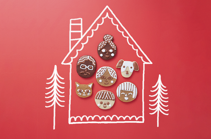 A group of cookies decorated to look like different faces sit on a red surface; drawn around the cookies is a simple illustration of a house and two evergreen trees.