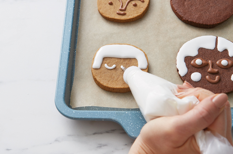 A woman holds a piping bag full of white royal icing and uses it to draw a face and hair on a round gingerbread cookie.