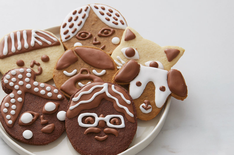 A white platter is filled with chocolate cookies, sugar cookies and gingerbread cookies decorated with white and chocolate royal icing to look like different people's and pet's faces.