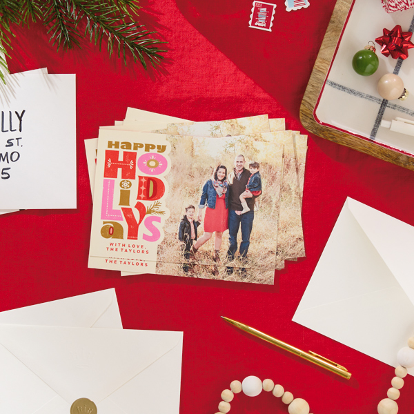 A stack of personalized holiday photo cards lies on a table covered with a bright red table cloth, surrounded by envelopes, a pen and holiday greenery.