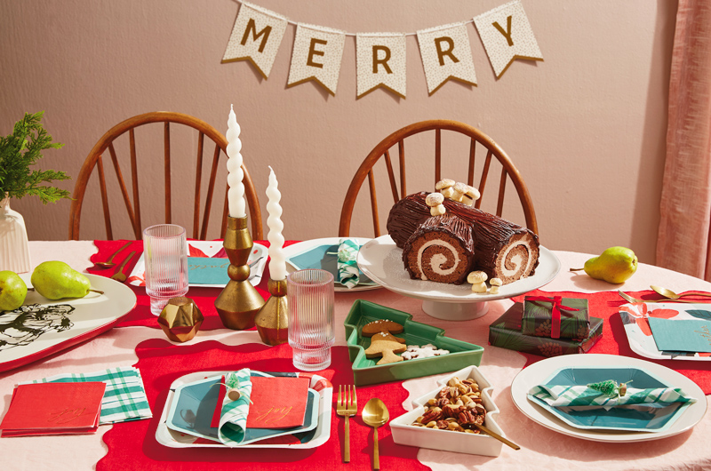 A Christmas tablescape featuring Hallmark Celebrate! partyware products like dark green hexagon-shaped plates and dark green and white plaid napkins; at the center of the table is a traditional yule log cake covered in chocolate frosting and meringue marshmallows, sitting on a simple white cake stand; nearby is a set of mod gold candle holders with white, twisted candles in them; behind the scene is a banner that says 