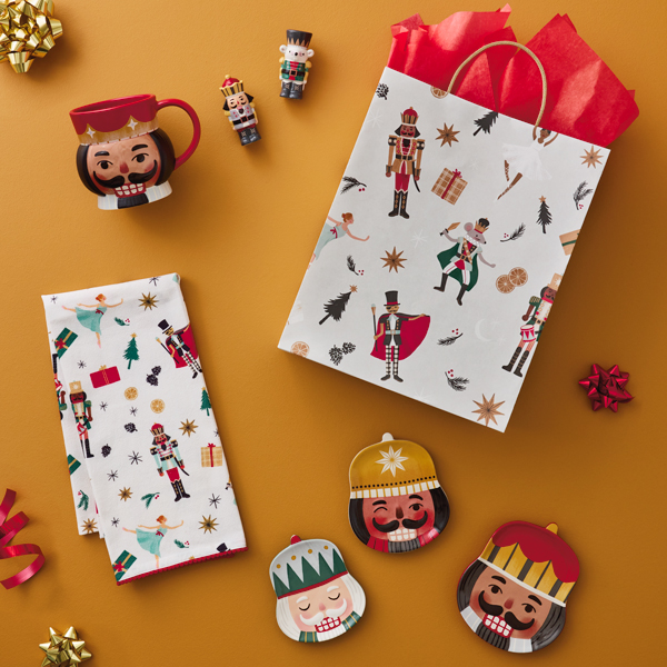 A collection of nutcracker-themed holiday gifts lay scattered on a deep yellow-gold surface; gift items include nesting dishes in the shapes of different nutcracker faces, a mug in the shape of a nutcracker's head, a nutcracker and mouse king salt and pepper shaker set, and a nutcracker ballet illustration tea towel; a gift bag featuring a nutcracker ballet illustrated pattern and stuffed with red tissue paper lays among the scattered gifts.