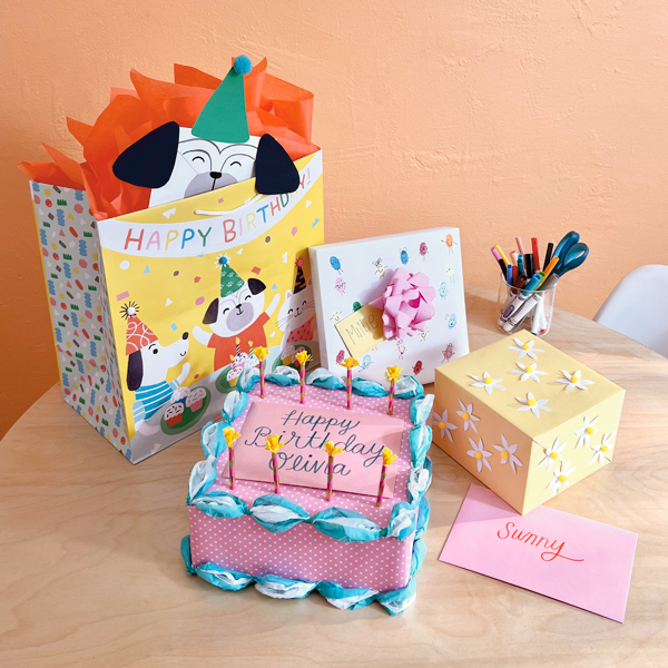 An assortment of gifts wrapped in wrapping paper and gift bags are decorated in fun and playful ways for kids, including a gift that's been wrapped and decorated to look like a birthday cake with candles, a gift covered in thumbprints that have been turned into little cartoon characters, a gift wrapped with yellow paper and decorated with 3D daisy flowers, and a gift in a bag with a matching card envelope that's been made to look like a dog wearing a party hat.