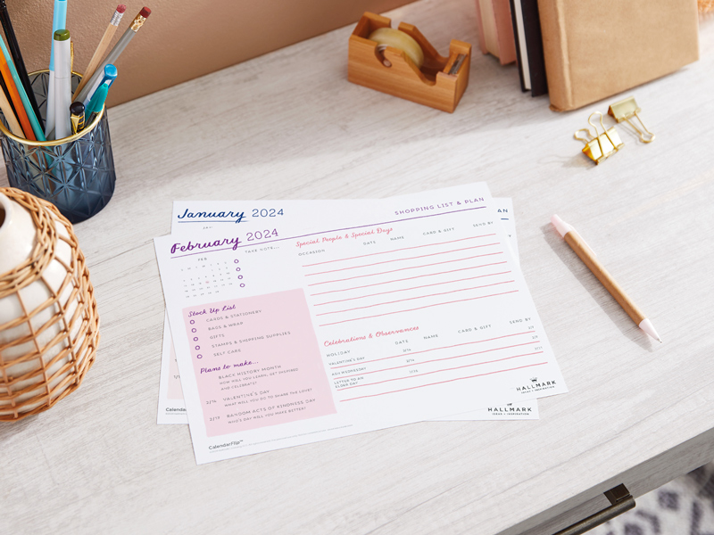 A printed February 2024 celebration planner page sitting on a home office desktop, surrounded by desk accessories.