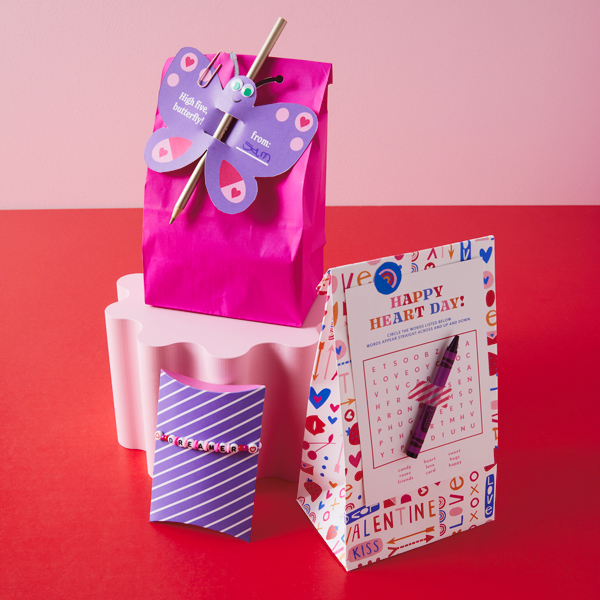 Three different DIY Valentine's Day treat bags sit assembled on a bright red surface; one is a purple pillow box filled with treats and embellished with a handmade, beaded friendship bracelet; another features our free printable Valentine's Day wordsearch and a Crayola crayon; the third and final treat bag sits on top of a light pink riser—it's a bright pink bag with one of our free printable Valentine's Day friends attached to it with a paper clip, with a colored pencil stuck through it.