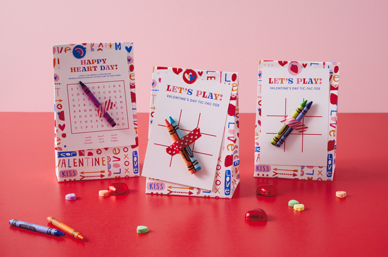 Valentine's Day treat bags featuring our free printable games and Crayola crayons sit assembled on a bright red surface that's scattered with red foil wrapped heart-shaped chocolates and candy conversation hearts.