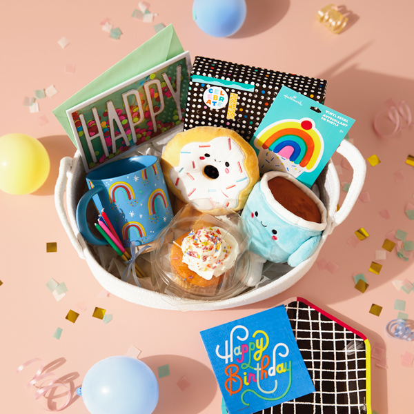 A birthday care package for a coworker or friend that includes a Coffee and Donut Better Together magnetic plush set, a light blue coffee mug with rainbows on it, a rainbow cupcake vinyl decal, tall, skinny candles, a cupcake with white frosting and rainbow-colored sprinkles, a birthday card with the word 