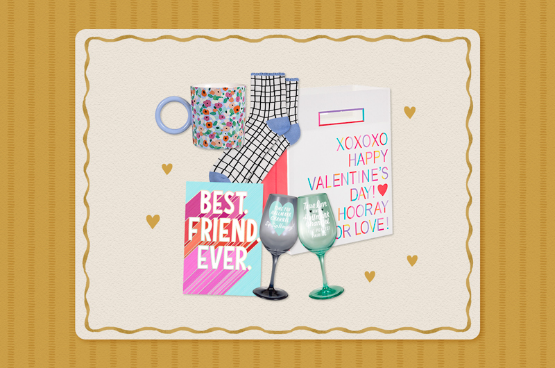 A collection of Valentine's Day gifts for a friend that includes a pair of socks with a black and white grid print and light blue toe and heel seams, a floral mug, a set of Hallmark Channel wine glasses, a card that reads, 
